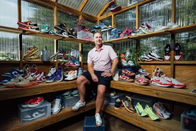 The image shows Jacob Barney wearing a light pink shirt and dark shorts sitting in a wooden greenhouse surrounded by an extensive collection of colorful Nike sneakers. He is smiling, looking off-camera. The shelves around him are filled with a variety of sneakers in different styles and colors, neatly arranged, with some potted plants adding greenery to the scene.